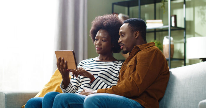 Young happy beautiful african american couple sitting at home on couch and browsing online catalog using tablet.